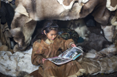 Nadia Takui, a Chukchi girl, dressed in a traditional reindeer skin Kamleika (parka), reads a magazine at the front of the polog (sleeping area) in a Yaranga (tent) at a reindeer herders' summer camp. Iultinsky District, Chukotka, Siberia, Russia
