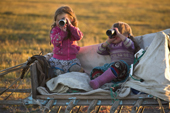 Sitting on a sled two Nenets girls, Darina Khudi & Angelina Laptander (right), play at spotting reindeer through binoculars at a reindeer herders' summer camp, Yamal Peninsula, NW Siberia, Russia